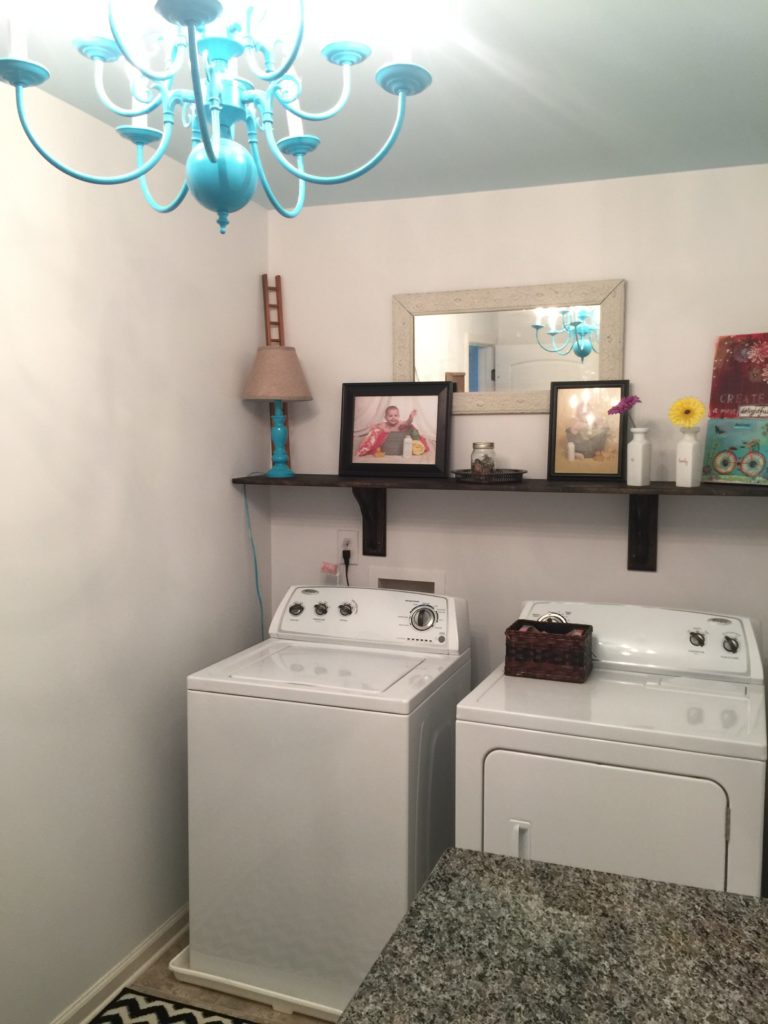 A laundry room with white walls, a blue ceiling, a blue chandelier, and a decorated shelf above the washer and dryer.