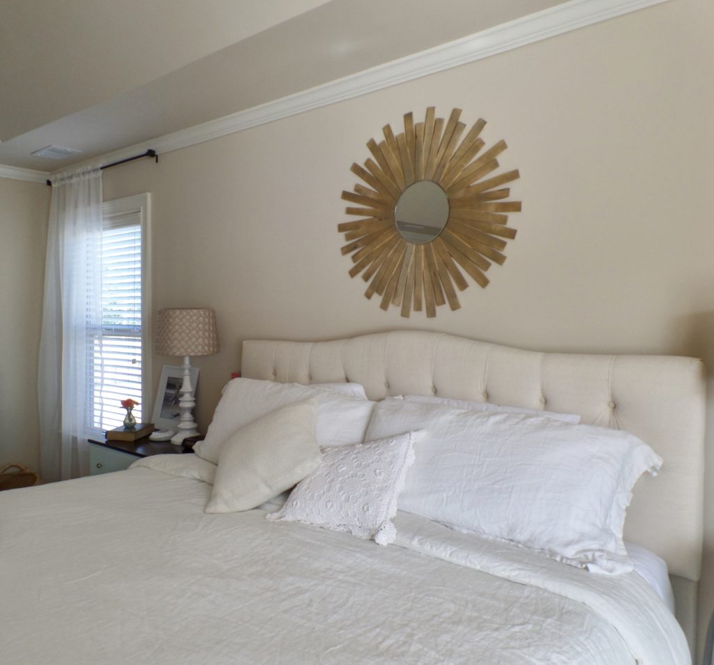 Before image of a master bedroom makeover with a large bed that has a cream-colored tufted headboard and white bedding, smooth cream walls, and a sunburst mirror over the bed.