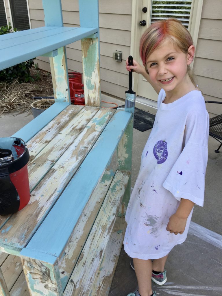 A child uses a paint brush to paint a wooden planter shelf with teal paint.