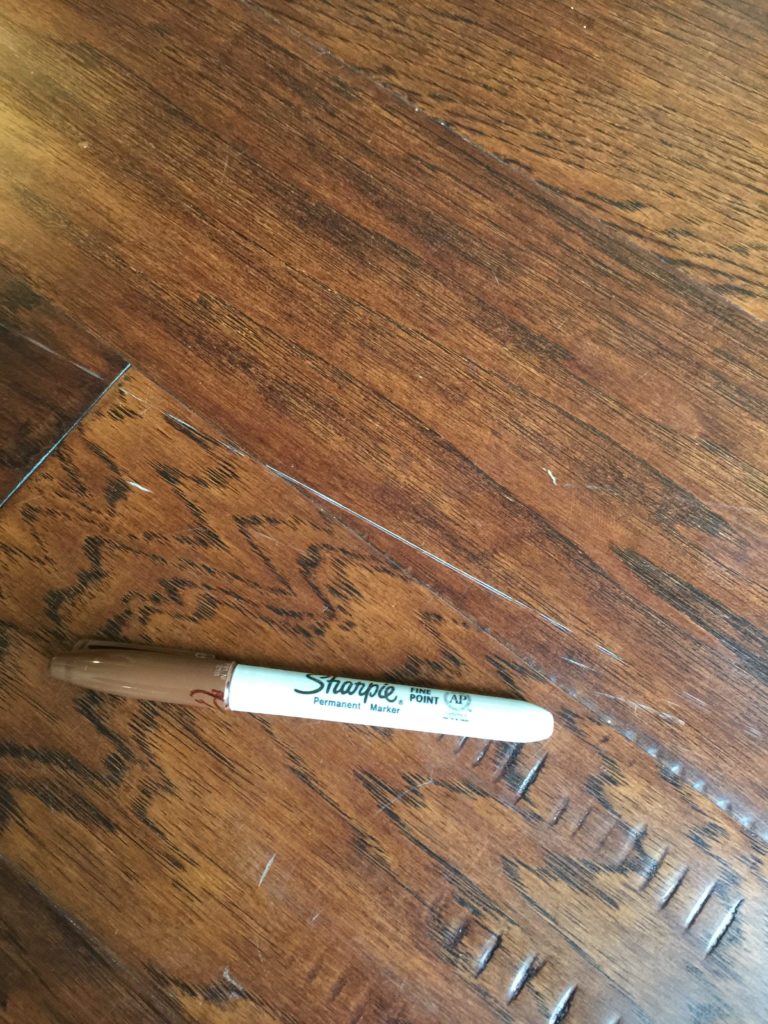 A brown sharpie marker next to a scratch on dark wood floors.