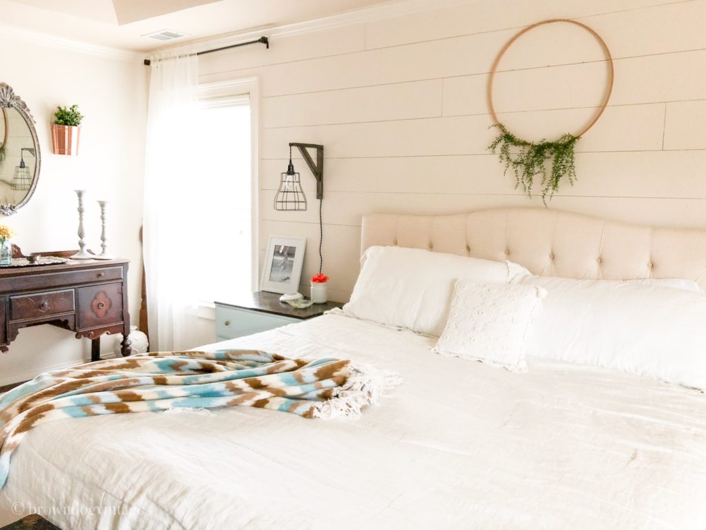 A tan and white bed with a tufted headboard next to a window with sheer white curtains.