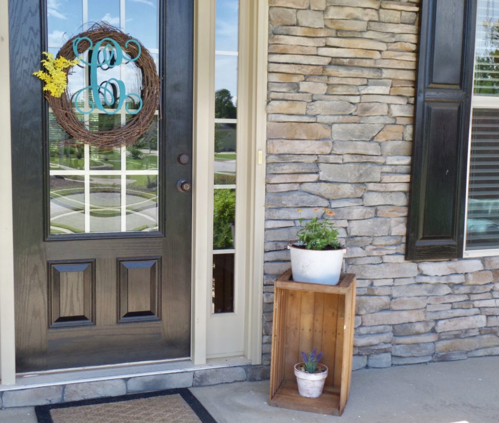 A wooden crate turned into a plant stand with two white pots with flowers inside next to a front door.