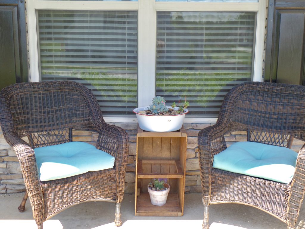 Two wicker chairs on a front porch with a wooden crate turned into a plant stand with two white pots with flowers inside.