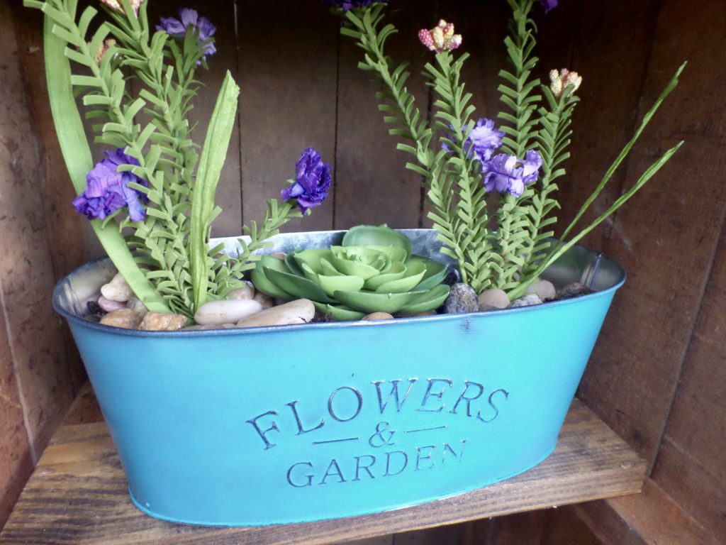 A teal metal planter that says \"Flowers & Garden\" on the front with flowers and succulents inside.