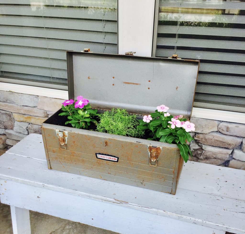 An old metal toolbox with flowers growing inside on a front porch.