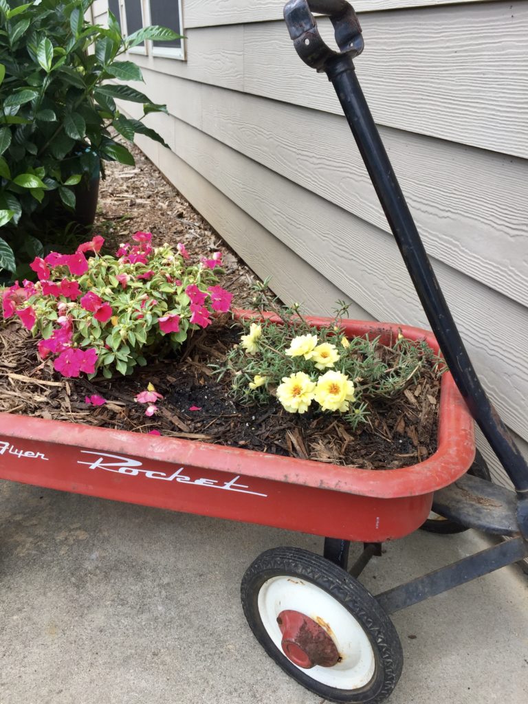 A little red wagon turned into a planter box with pink and yellow flowers growing inside.