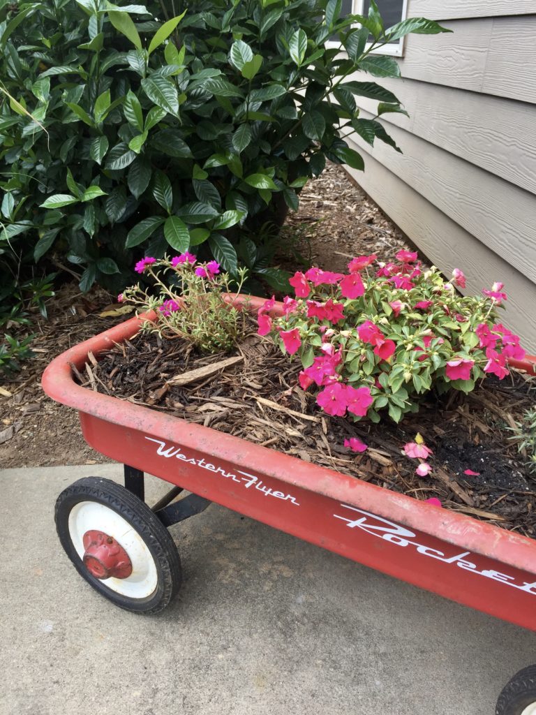 A small rusty red wagon turned into a planter with pink flowers growing inside.