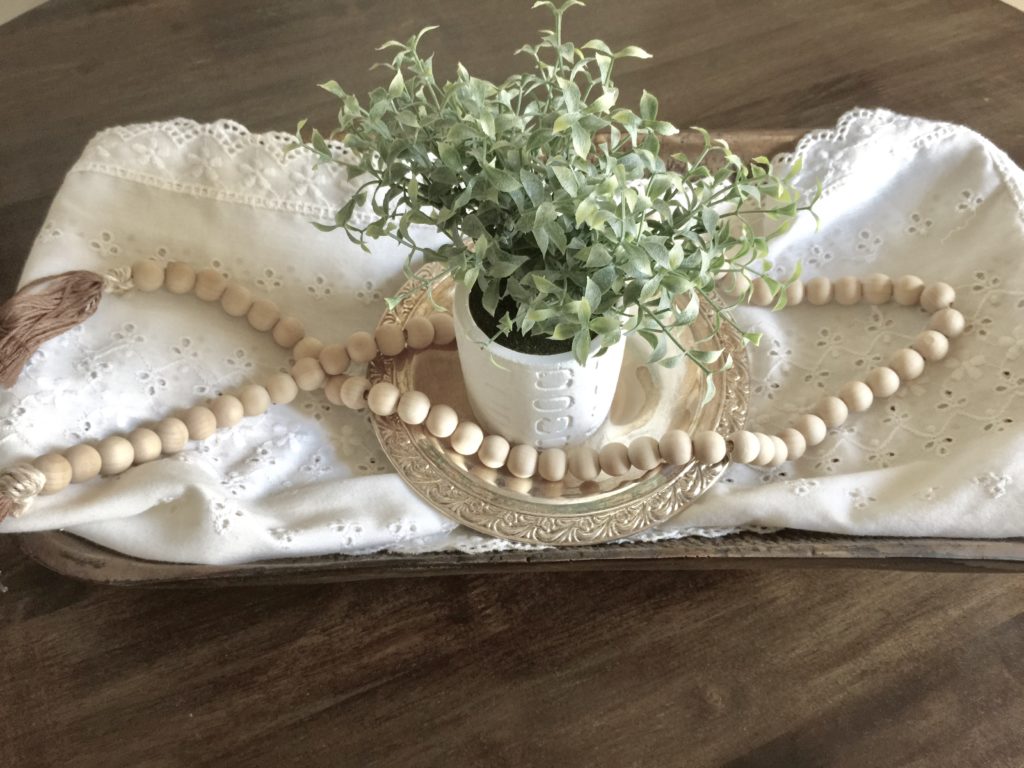 Wooden bead garland on a table with a white cloth and greenery in a small white pot.