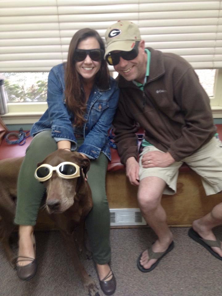 A man and a woman pose with their dog, all wearing sunglasses.