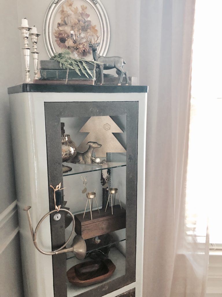 An antique medical cabinet in the corner of a room with gold Christmas decorations inside and on top.