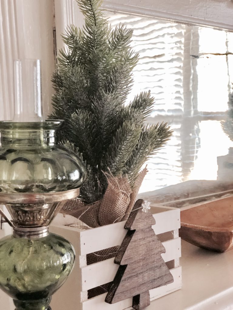 A mini fir tree in a burlap sack inside a wooden crate on a fireplace mantel.