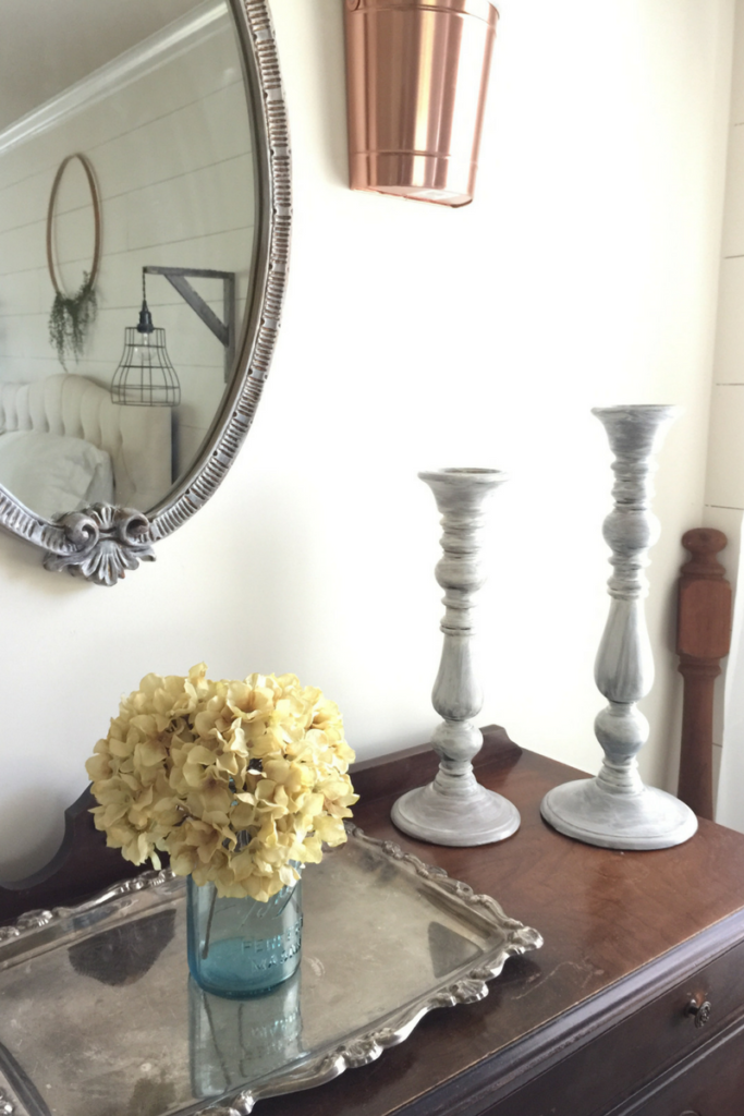 Candle sticks and a glass vase with yellow flowers on a dark wood side board under an antique silver mirror.