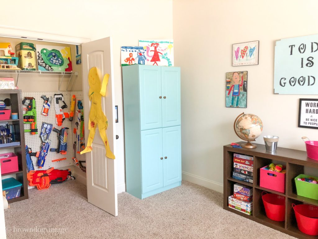 Nerf guns hang from a storage system in a playroom closet next to a blue cabinet and dark brown cube storage shelves.