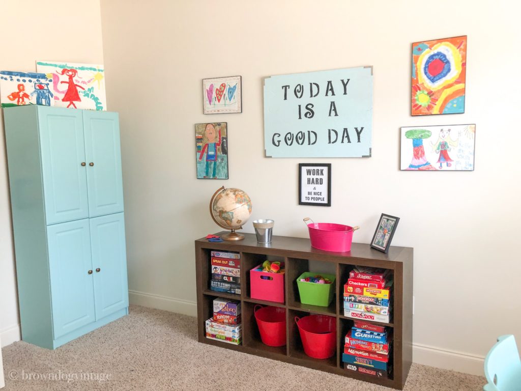 A tidy playroom with a storage cabinet and shelves and artwork on the walls including a playroom sign that reads \"Today is a Good Day\".