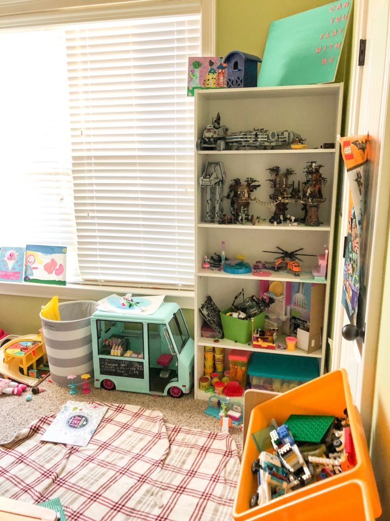 A children\'s playroom with a tall shelf, lots of toys, and a large window.