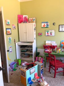 A cluttered playroom with a white cabinet, green desk, and a red chair.