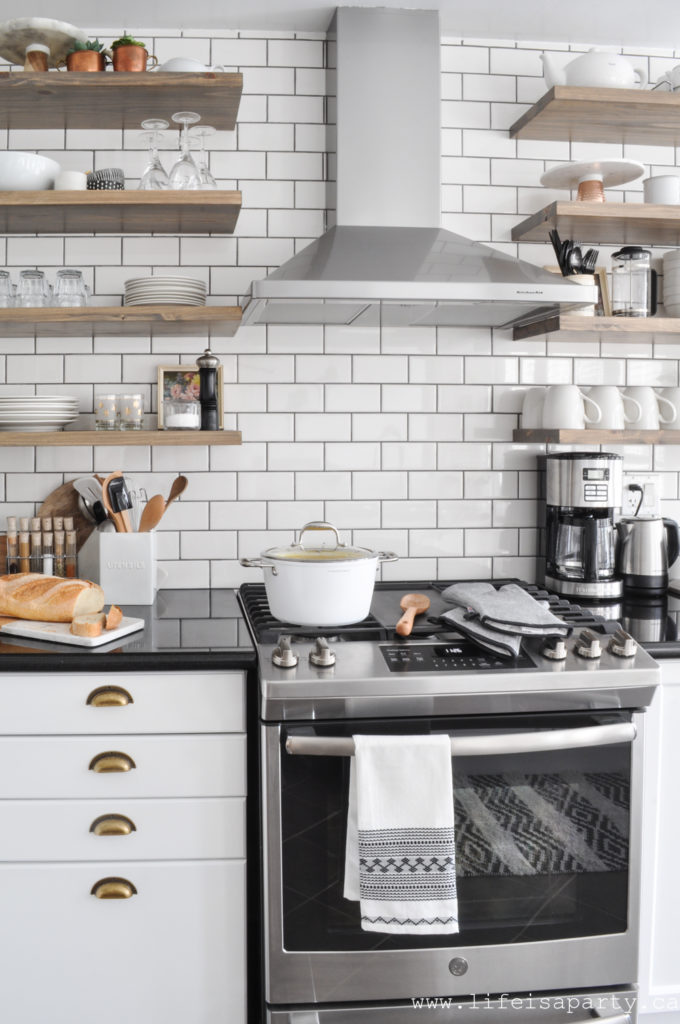 A kitchen with white cabinets, a black countertop, a metal vent hood over the stove, floating wooden shelves, and a white subway tile backsplash.