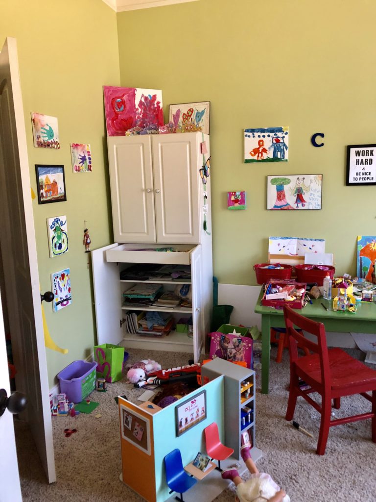 Cluttered playroom with a white storage cabinet on the back wall and a dollhouse in the middle of the floor.