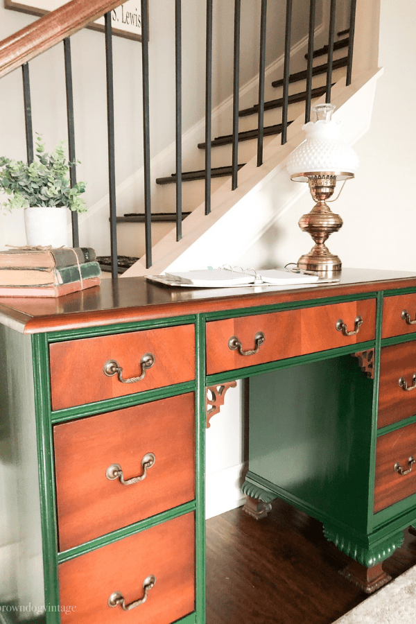 An antique green and wood desk with a brass lamp, books, and a faux potted plant on top.