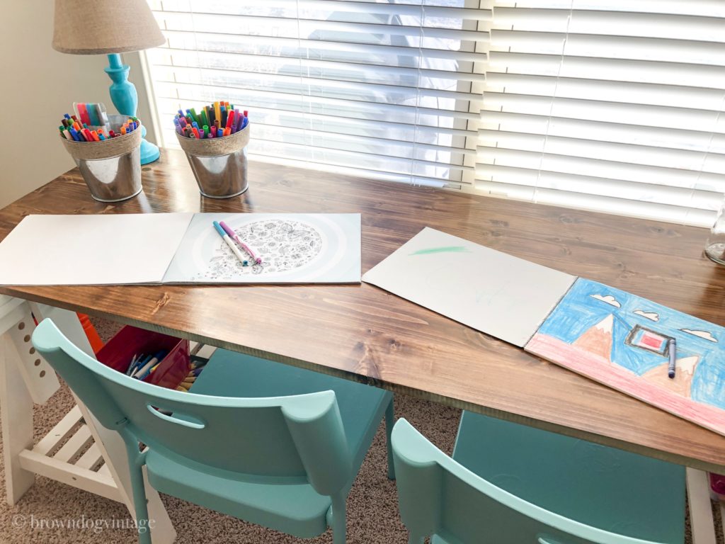 A children\'s desk with coloring books and crayons on top next to a window with partially opened blinds.