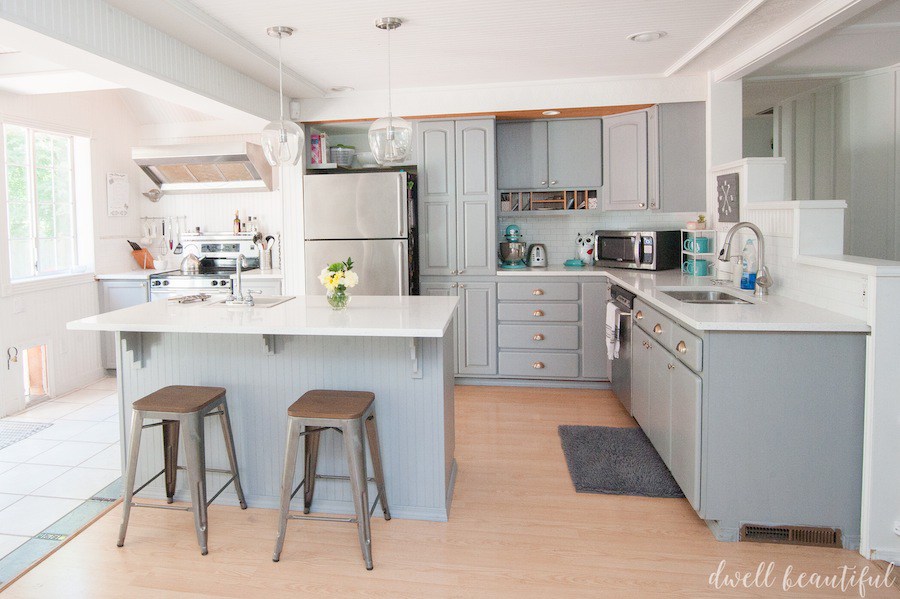 A kitchen with gray cabinets and white countertops with an island and metal barstools. 