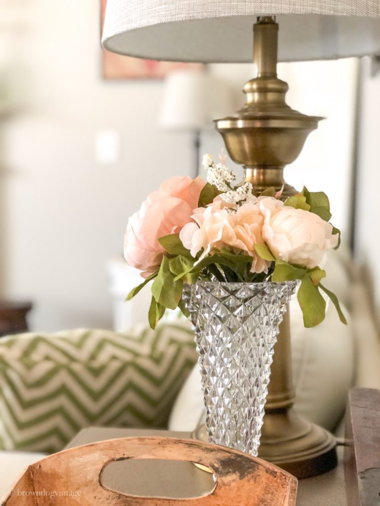 Pink faux flowers in a vintage glass vase next to a lamp on an end table.