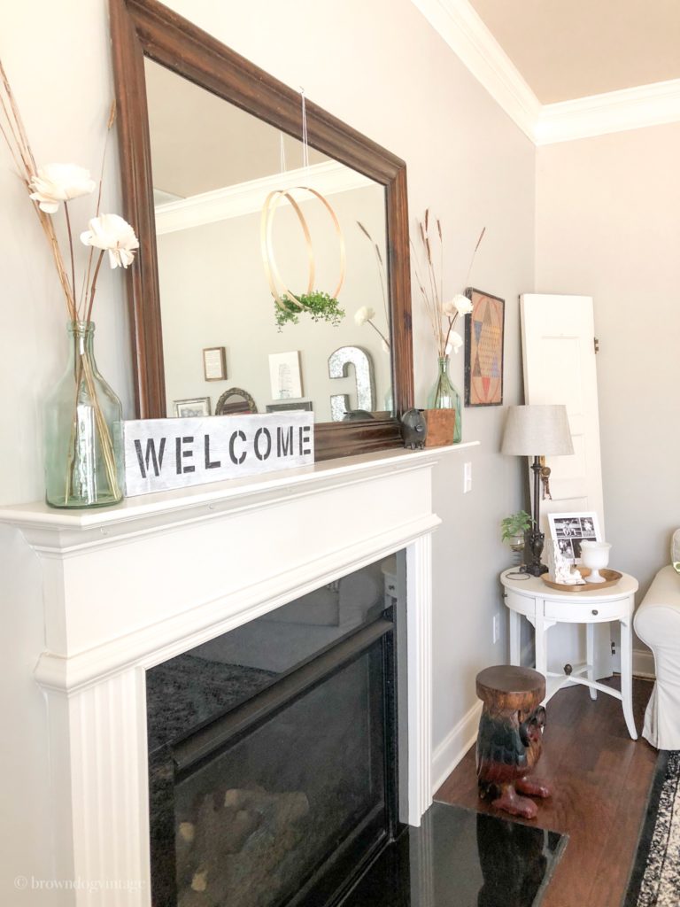 White flowers in a green glass vase and a welcome sign on a fireplace mantle.