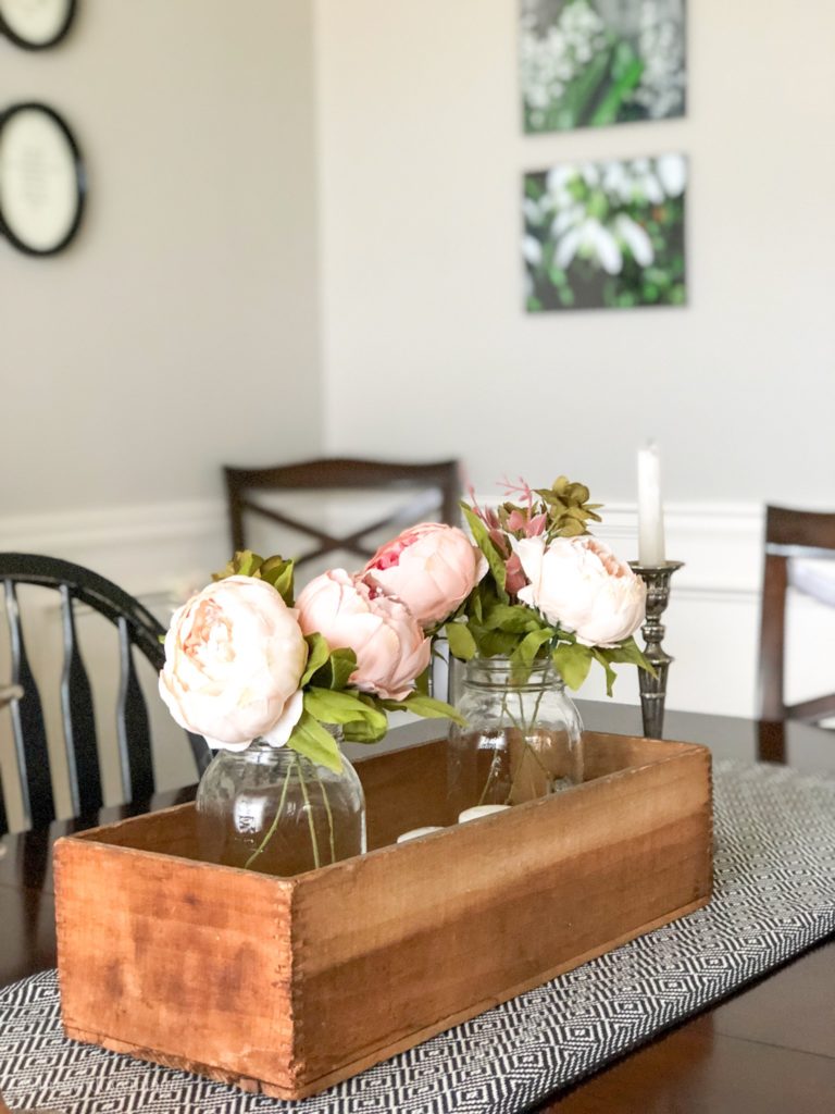 Faux pink peonies in two glass jars in a wooden box on a dining room table.