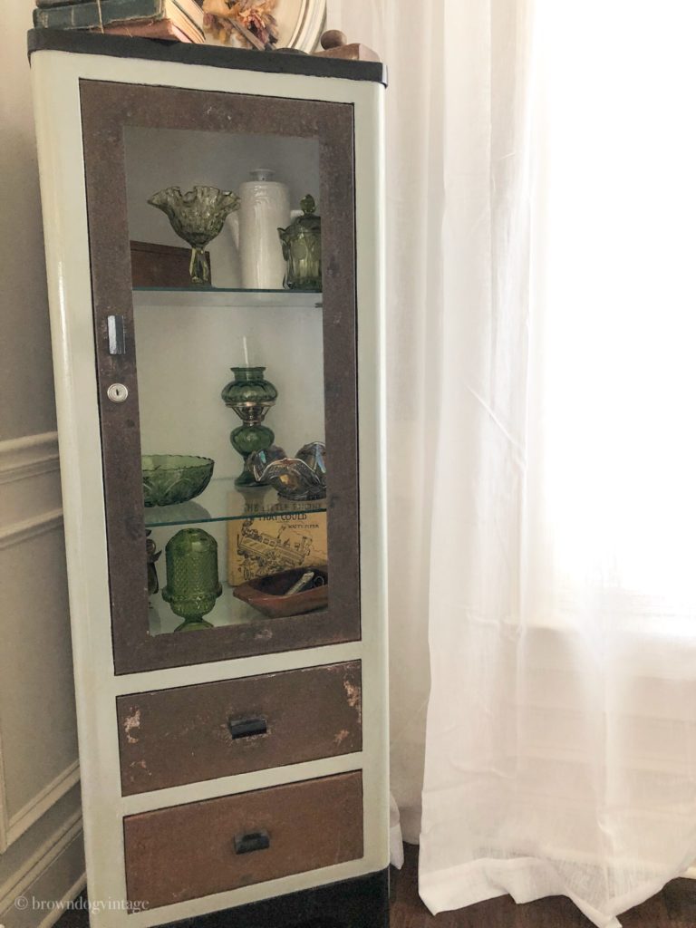 A corner curio cabinet with decor inside next to a window with sheer white curtains.