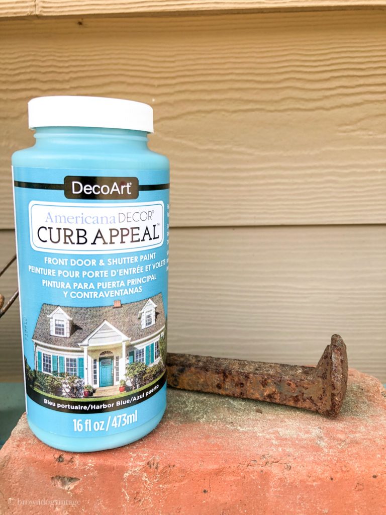 A bottle of blue Curb Appeal DecoArt paint sitting next to a rusty piece of metal by the side of a house outside.