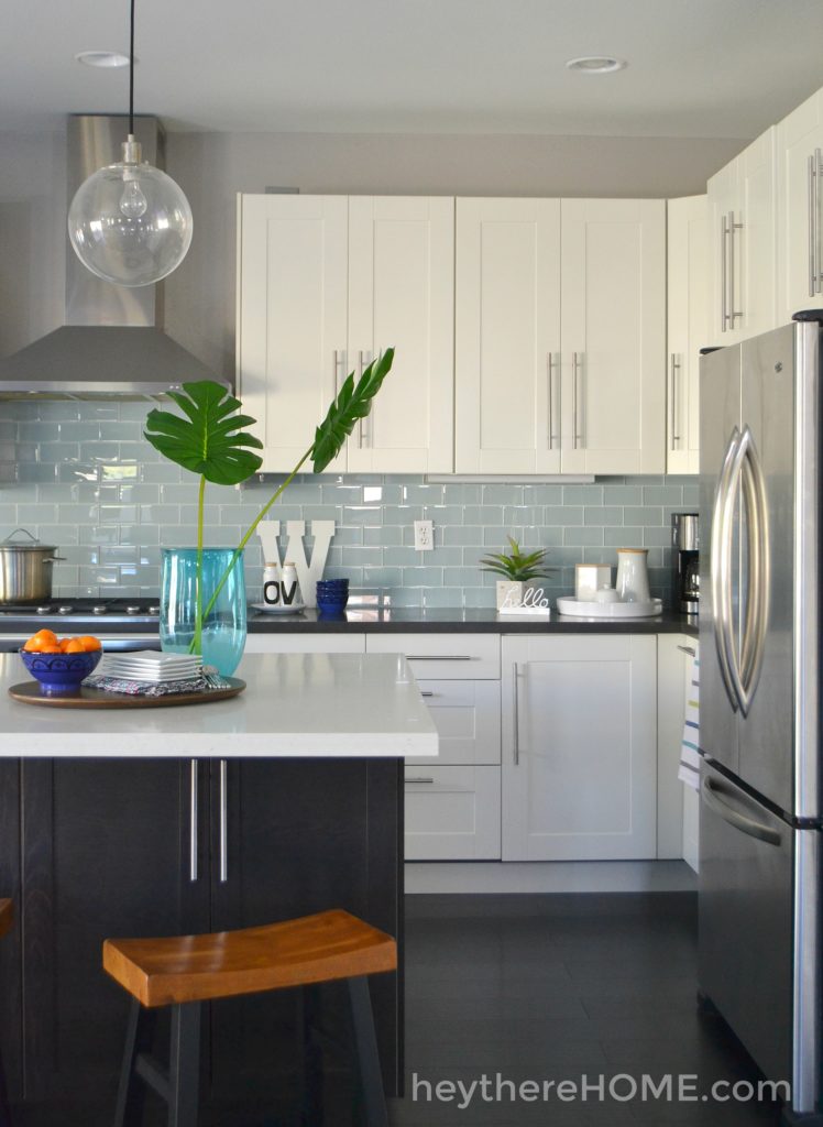 A kitchen with white cabinets and teal backsplash with dark countertops and a black island with white countertops.