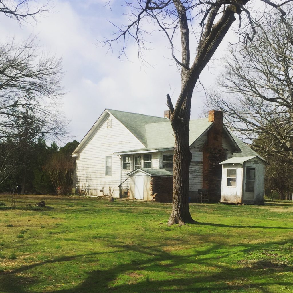 A Farmhouse with a tall tree in front without any leaves and a large green yard.