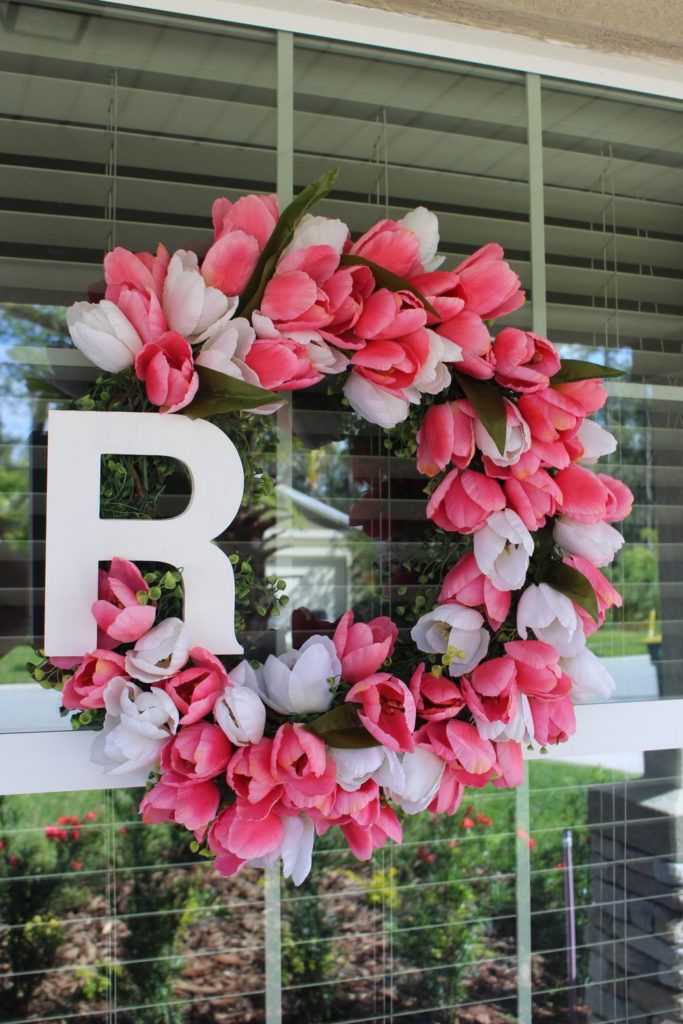 A homemade tulip wreath made from faux pink and white tulips with a wooden letter R hangs on the window of a house.