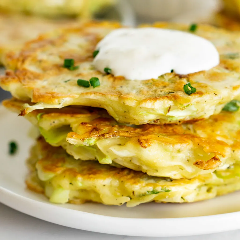 A stack of cabbage fritters on a white plate with green garnish and white cream on top.