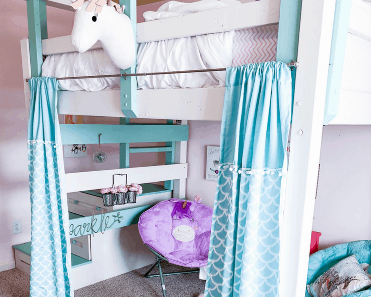 A bunk bed with space underneath for chairs decorated with a unicorn head and blue curtains.