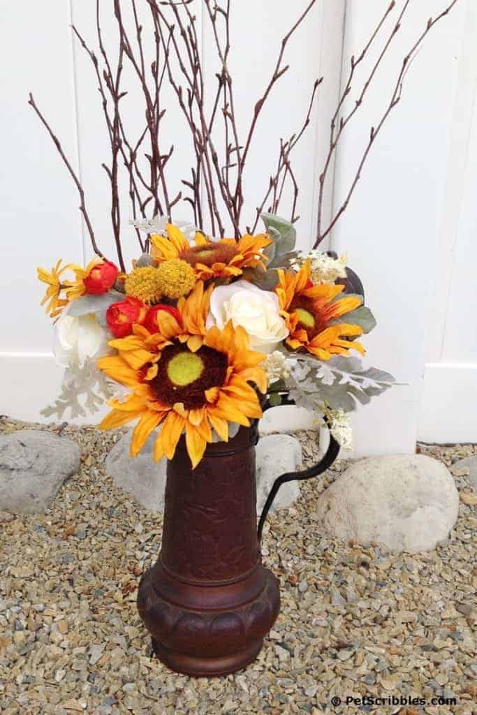 Sunflowers and fall foliage in a rustic container sitting on gravel in front of a white fence.