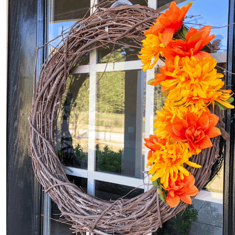 A stick wreath with red and orange flowers decorating one side, hanging from a front door.