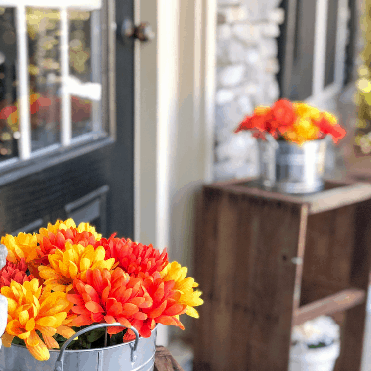 A front porch with two tables on either side of the front door and metal containers with red, yellow, and orange flowers inside.