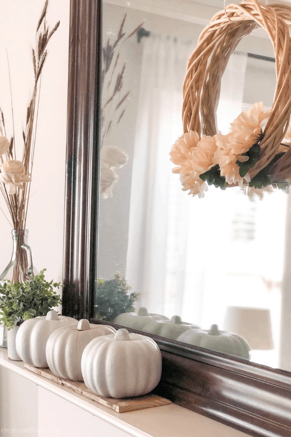 A wreath with pale orange flowers hangs over the mirror of a fireplace mantle with white pumpkins.