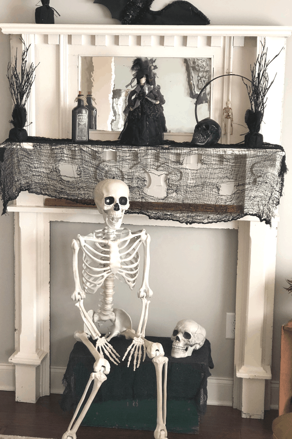 A skeleton sits in front of a fireplace mantle covered in a holey black cloth with black Halloween decor.