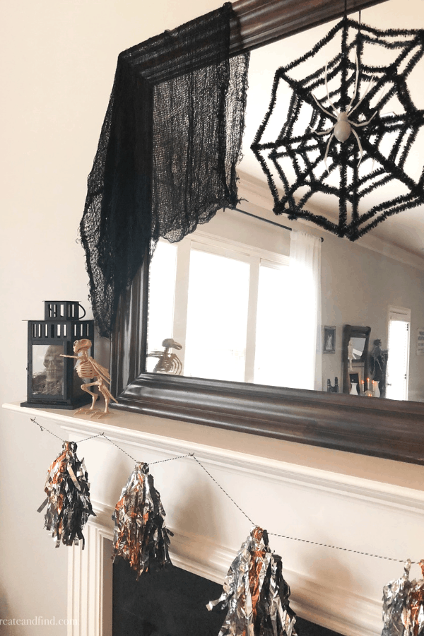 Spooky decorations on a fireplace mantle and mirror, including a spider web and orange and black tassel garland.