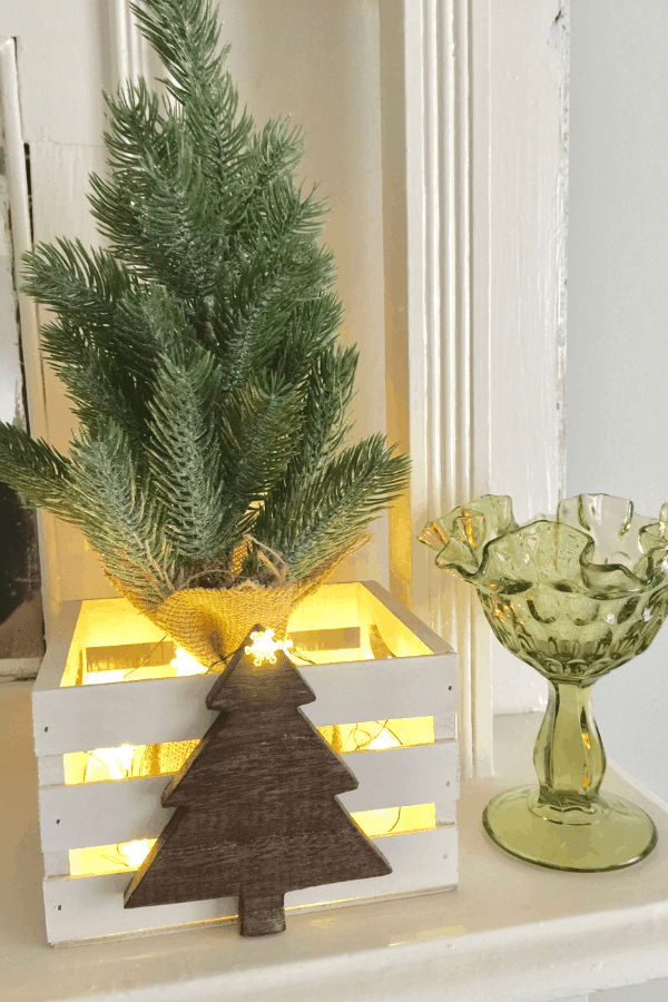 A miniature faux Christmas tree in a wooden create on a fireplace mantle.