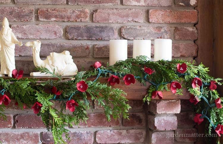 Green garland with red flowers draped over a brick fireplace mantle with three white candles and a nativity scene.