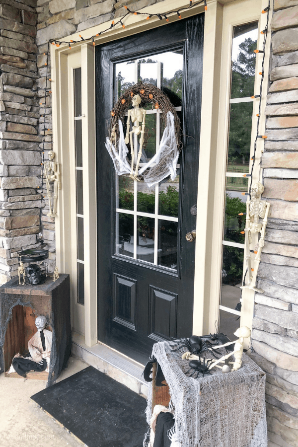 A wreath with a miniature skeleton and cobwebs hangs from the front door of a house with spooky decor on the stoop.