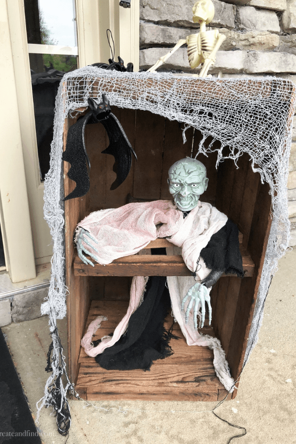 A green ghoulish skeleton in a wooden crate draped with gauze on a front porch.