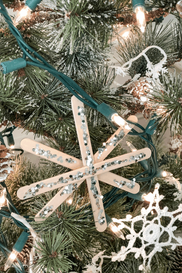 A snowflake made out of popsicles sticks and glitter hanging on a Christmas tree.