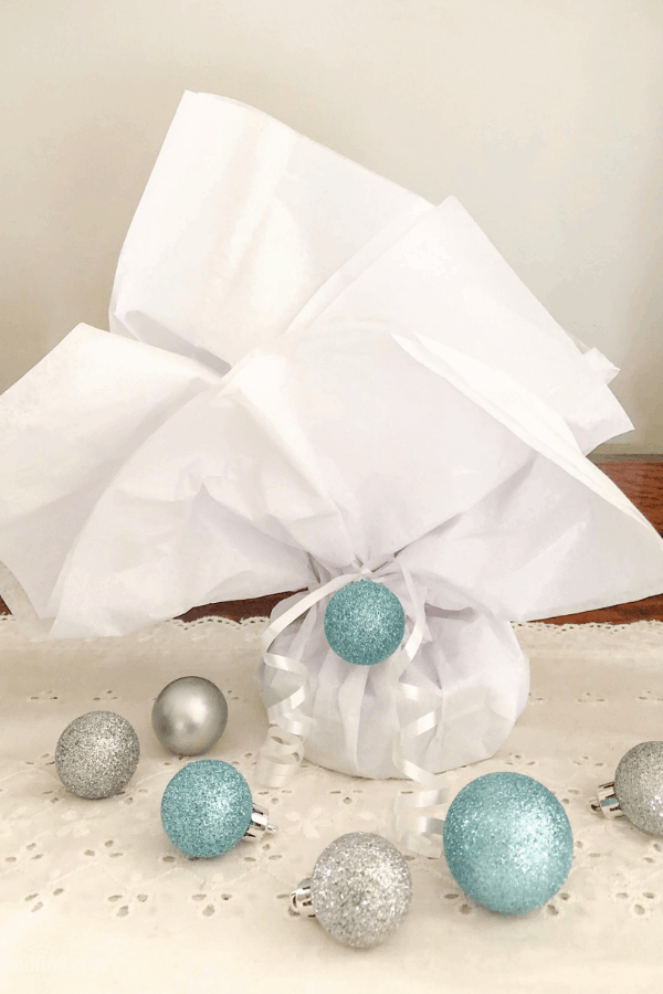 A small gift wrapped in whit tissue paper with a white ribbon and glittery blue ornament.