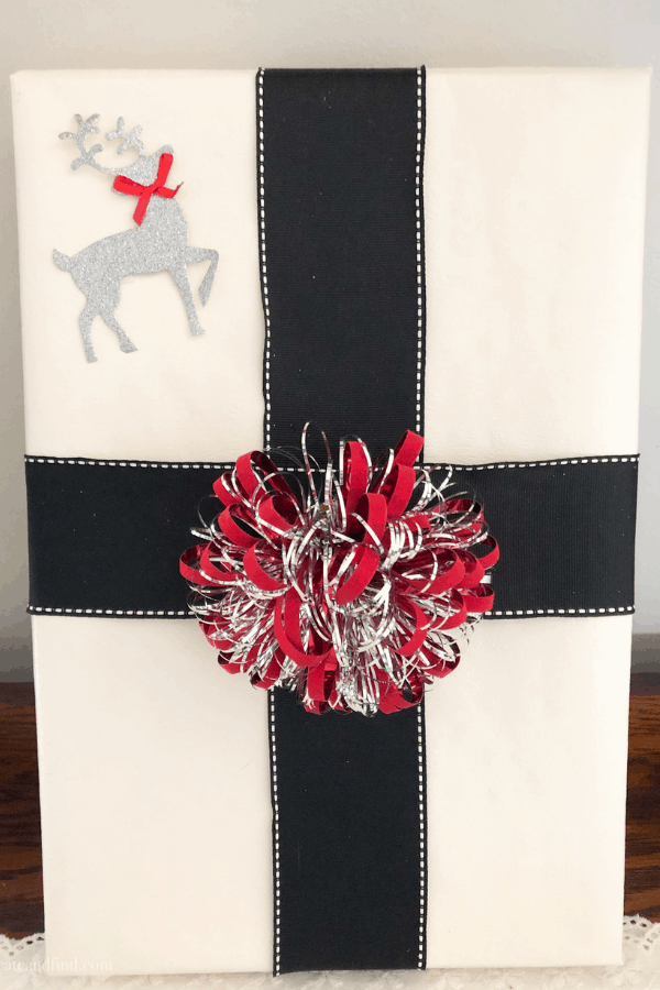 Close up of a Christmas present wrapped in white paper with black ribbon and a silver and red bow and reindeer cutout.