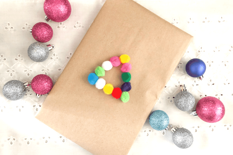 A rectangular Christmas gift wrapped in brown paper and decorated with colorful felt balls in the shape of a triangle.