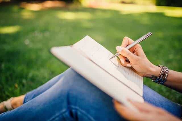 A woman writes in a journal while sitting outside on the grass.